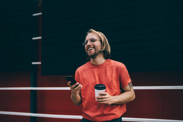 Happy laughing bearded modern male with long hair in red t-shirt watching on mobile phone holding cup of takeaway coffee in street - 写真・画像
