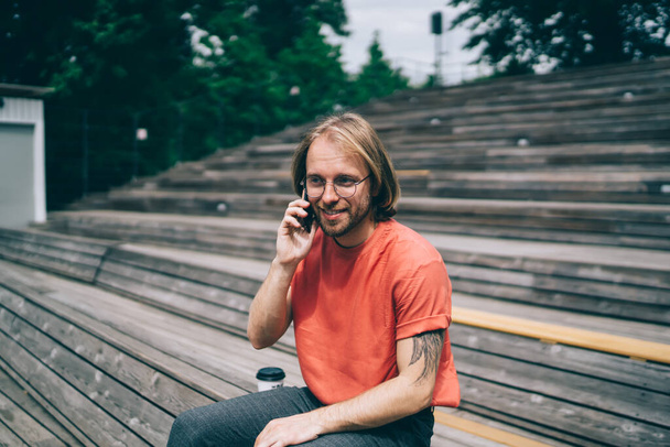 Smiling bearded modern hipster with tattoo in glasses  with long hair in red t-shirt calling on mobile phone in city street  - Фото, изображение