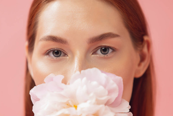 Close-up portrait of beautiful redheaded woman with flower isolated over pink studio back ground. Nude color, diet, cosmetics, natural beauty and aesthetic cosmetology concept. - Photo, image