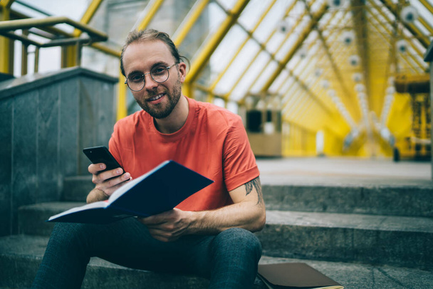Cheerful smiling male in casual clothes and glasses using phone reading notepad sitting on stairs in pedestrian bridge looking at camera - Zdjęcie, obraz