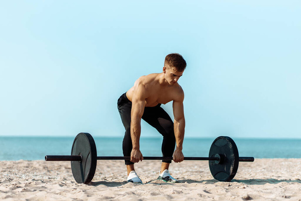Healthy handsome active man with fit muscular body, young muscular man lifting weights, doing exercises, Sporty man exercising on the beach, Outdoor training, Sport and fitness concept - Foto, Bild