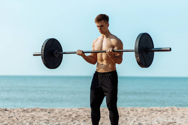 Muscular guy bodybuilder doing exercises with big dumbbells outdoors on the beach, on a summer sunny day, training on the sea coast on the background - Foto, Imagem