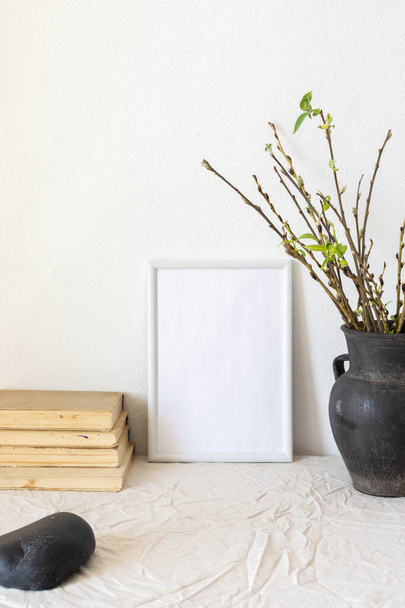 Home decor mock-up, blank picture frame near white painted concrete wall , black earthenware jug with pussy willow branches and blossoming green leaves, old books - Foto, immagini