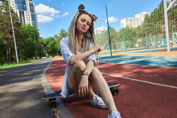 A teenage girl with long pigtails sits on a longboard near a sports ground - Foto, Imagen