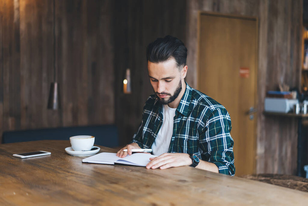 Caucaisan male writer reading informative notes in education textbook spending time in local cafeteria, intelligent hipster guy in shirt creating publication ideas using notepad in coffee shop - Foto, Bild