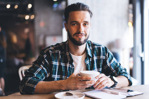 Portrait of happy male writer with textbook smiling at camera spending daytime for creating idea in cafe interior, successful hipster guy with coffee cup sitting at cafeteria table and posing - Photo, image