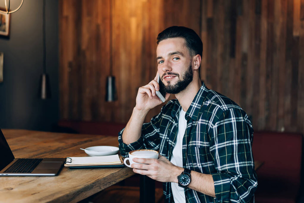 Portrait of Caucaisan male student with cappuccino cup in hand using modern cellphone gadget calling and talking, young man with coffee looking at camera while making smartphone contact conversation - Foto, Imagem