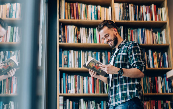 Side view of cheerful male student in casual clothing reading best seller novel in public bookstore, smiling hipster guy enjoying time in library choosing literature during leisure with hobby - Фото, зображення