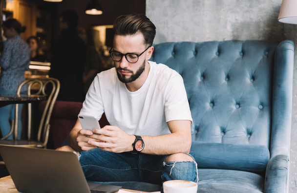 Millennial blogger in spectacles using bluetooth connection on smartphone and laptop technologies for sharing media files during freelance remote working, man checking received cellular notification - Fotografie, Obrázek