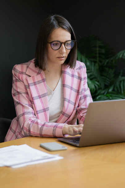 Femme d'affaires européenne confiante dans une veste rose et avec un ordinateur portable dans son bureau de travail - Photo, image