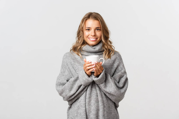 Image of beautiful smiling woman drinking coffee or tea from white mug, standing over white background - Foto, Imagem
