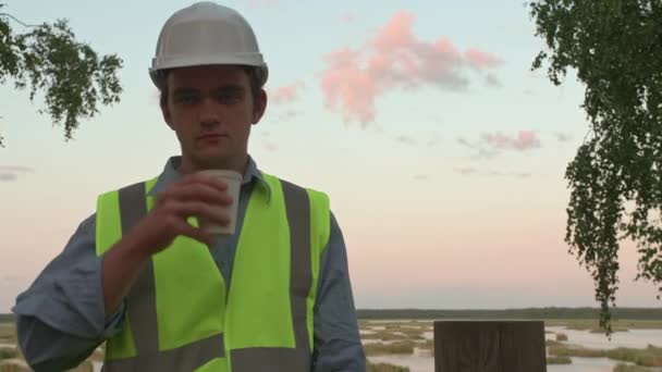 Jeune écologiste constructeur fatigué de boire du café et regardant la caméra, il est vêtu d'un jaune réfléchissant et d'un casque blanc, le concept d'une catastrophe écologique et sa solution - Séquence, vidéo