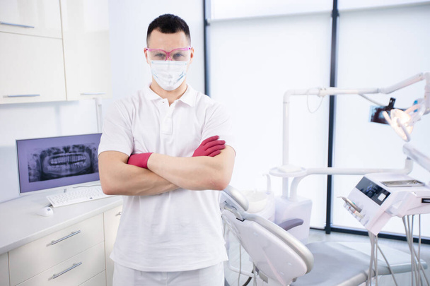 Πορτρέτο ενός νεαρού οδοντίατρου με προστατευτική μάσκα, γυαλιά εργασίας και με λευκή στολή στέκεται στο γραφείο κατά τη διάρκεια της καραντίνας του κορωναϊού. Επείγουσα περίθαλψη - Φωτογραφία, εικόνα