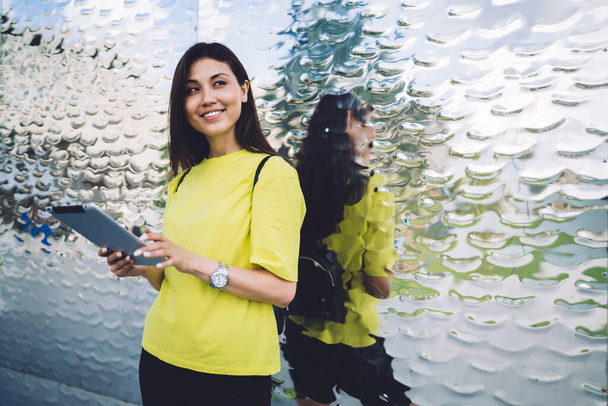 Boldog etnikai nő sportruházatban és hátizsákban áll üvegfal mellett táblagéppel, miközben mosolyog és félrenéz az utcán - Fotó, kép