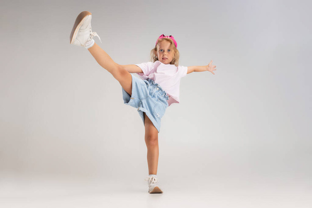 Beyaz stüdyo arka planında izole bir şekilde tek bacağının üzerinde duran günlük giysiler içinde küçük beyaz bir anaokulu kızı portresi.. - Fotoğraf, Görsel