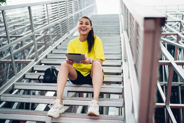 Zdola veselá etnická žena v neformálním oblečení s batohem sedícím na schodech v parku a usmívajícím se při prohlížení tabletu a při pohledu do kamery - Fotografie, Obrázek
