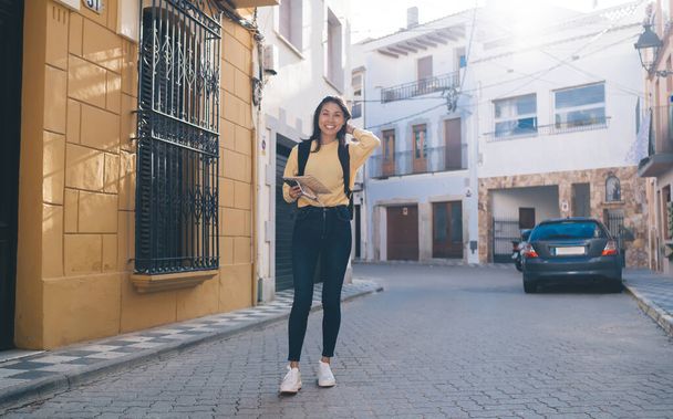 Full length portrait of cheerful Asian tourist smiling at camera during travel experience using paper location map for orientation navigation during solo vacations, happy hipster girl enjoying trip - Foto, imagen