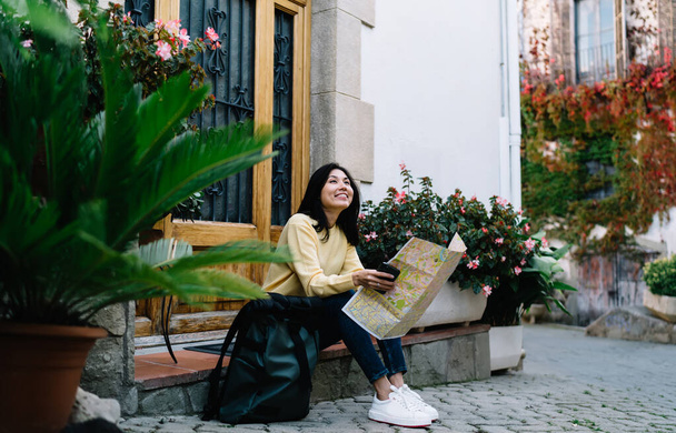 Carefree female tourist 20s with location map resting in amazing village sitting near building and dreaming, happy hipster girl enjoying summer vacations for recreating during getaway journey - Zdjęcie, obraz