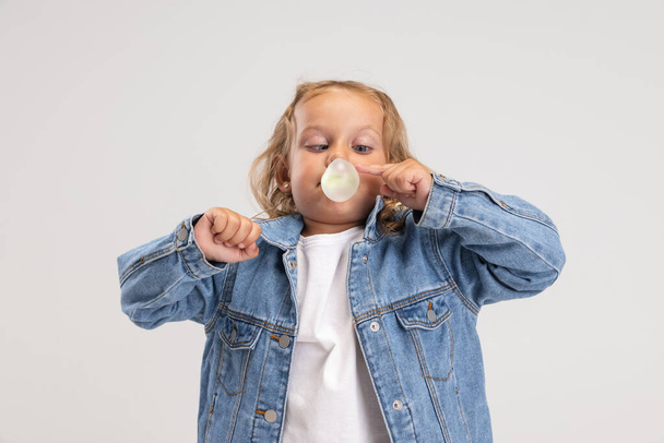 Одна маленькая дошкольная белая девочка в джинсовой одежде, раздувающая пузырь жвачки на белом фоне студии. - Фото, изображение