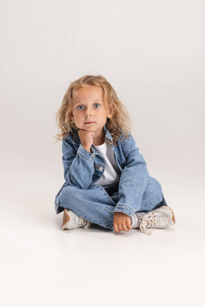 Портрет белой девочки в джинсовой одежде, сидящей на полу на белом фоне студии. - Фото, изображение