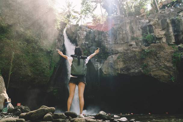 From below back view of full body unrecognizable female traveler with backpack enjoying picturesque waterfall with phone in hand while spending time in nature - Photo, Image