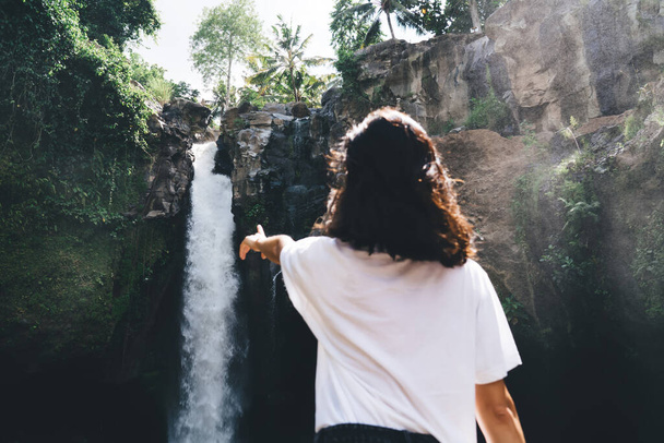 Back view of anonymous female traveler in summer wear enjoying waterfall among cliffs while spending time alone in tropical resort in Bali - Valokuva, kuva