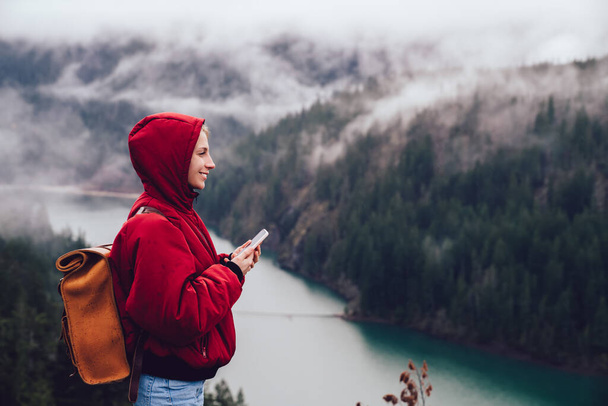Side view of happy female tourist with backpack and warm coat with hood using modern smartphone while exploring nature landscape with foggy coniferous forest and river in United States - Photo, image