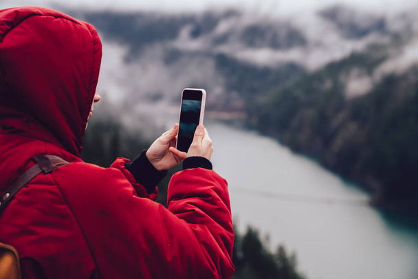 Back view of unrecognizable tourist in warm coat with hood standing on viewpoint and taking photo of amazing landscape with river in United States - Fotoğraf, Görsel