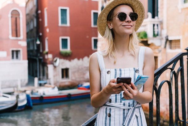 Young blond smiling woman tourist in dress and hat using mobile phone and map for planning trip in Venice Italy - Fotó, kép