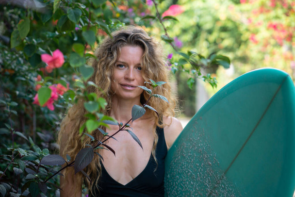 Portrait of young woman with blond long curly hair in bikini with blue surfboard on the street with tropical pink flowers and green bushes in Bali - Φωτογραφία, εικόνα
