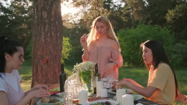 Grupa trzech młodych kobiet spędzających cudowny czas na świeżym powietrzu jedząc kolację pod drzewem w parku, materiał filmowy slow motion - Materiał filmowy, wideo