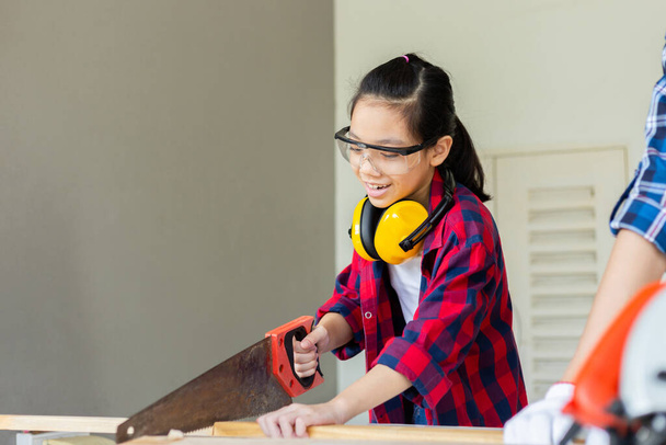 Çocuk marangozluk atölyesinde marangozluk atölyesinde ses azaltma kulaklıklarıyla ayakta duran Asyalı kız testereli ahşap işçiliği yapan bir marangoz. - Fotoğraf, Görsel