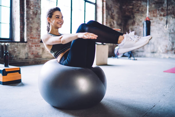 Full length of slim cheerful female in sportswear doing double crunches exercise on fit ball while having workout in spacious gym - Fotoğraf, Görsel