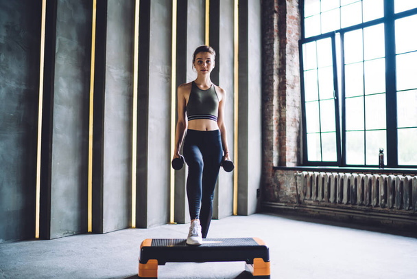 Determined caucasian woman in active wear prepare for cardio training in gym using equipment,serious beautiful female using dumbells and step for workout on slimming and muscles strength - 写真・画像