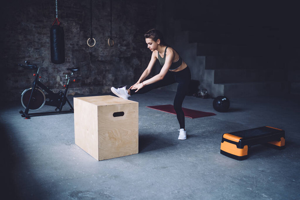 足を木製の箱の上に置く若い女性のフィット感の完全な長辺ビューの後に緊張した筋肉を伸ばすためのステップエアロビクストレーニング - 写真・画像