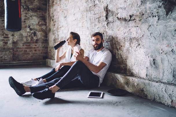 Side view of concentrated young bearded sportsman relaxing on floor in shabby gym after workout with girlfriend drinking water - Fotografie, Obrázek