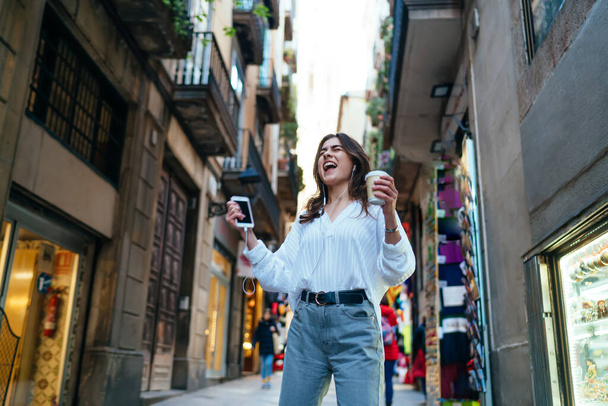 Happy Caucasian girl in electronic headphones enjoying touristic sightseeing singing while listening favourite audio song, excited female with takeaway coffee and cellular rejoicing while dancing - Photo, image