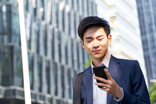 young asian businessman looking at messages on cellphone while walking in the street in downtown of modern city - Photo, Image