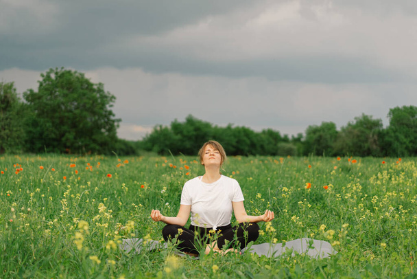 Woman in a white T-shirt and leggings doing yoga in a beautiful field on a sports mat. lifestyle and mindfulness concept - Foto, Imagen