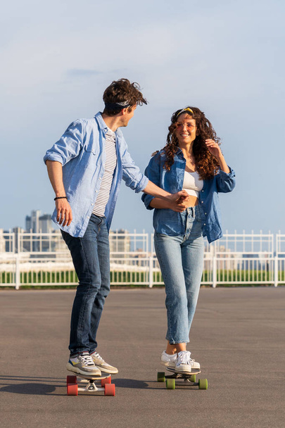 Stylish couple skateboarding together over urban skyline. Young adult man and woman on londboard - Foto, Imagen