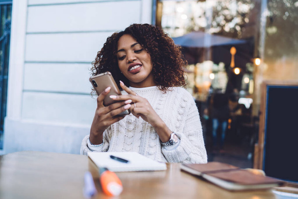 Smiling dark skinned curly hipster girl enjoying 4G connection for watching video on smartphone outdoors, positive african american millennial woman making book on web pages via mobile phone - Fotoğraf, Görsel