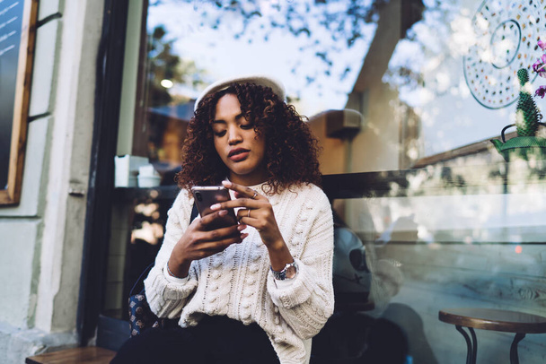 Przemyślana Afroamerykanka w luźnych ciuchach i kapeluszu siedząca na drewnianej ławce i przeglądająca smartfona podczas odpoczynku na ulicy - Zdjęcie, obraz