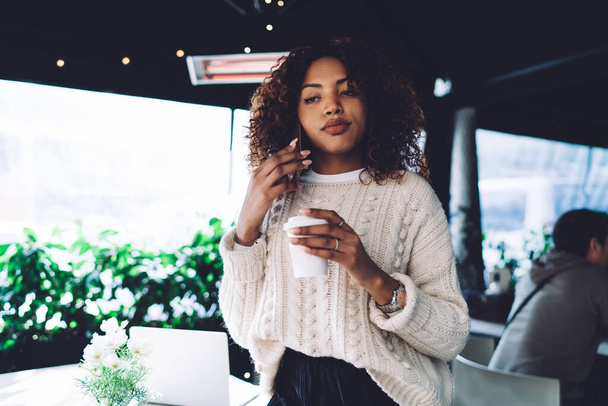 Serious African American female with curly hair wearing sweater making call on mobile while holding takeaway coffee at cafe terrace - Фото, зображення
