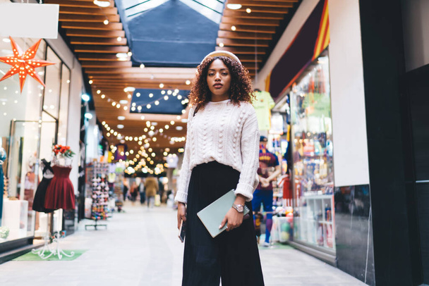 Half length portrait of trendy dressed african american female standing in shopping mall during free time, attractive 20s dark skinned woman dressed in stylish outfit posing in store looking at camera - Photo, Image