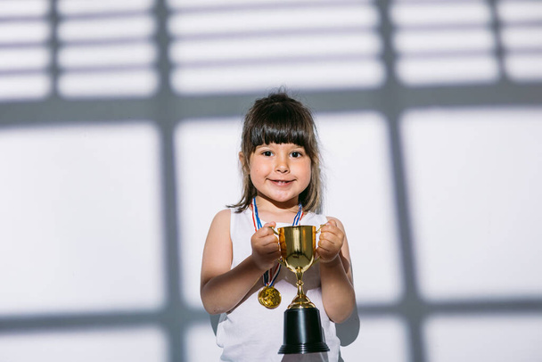 Tmavovlasá dívka se sportovními medailemi a pohárem trofejí, stojící ve stínu okna se záclonou. Koncept sportu a vítězství - Fotografie, Obrázek