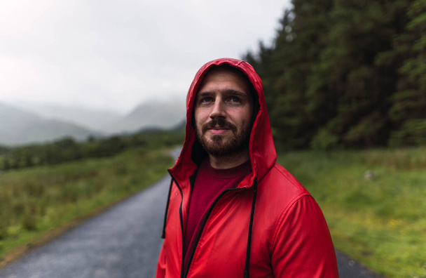 İskoçya, İskoçya 'da bulutlu bir günde bir ormanla çevrili boş bir yolda yürüyen kırmızı yağmurluklu bir adam. - Fotoğraf, Görsel