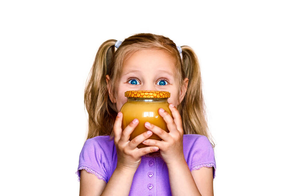 Klein meisje met een pot honing voor haar gezicht. Het kind op een witte achtergrond houdt een pot honing vast - Foto, afbeelding