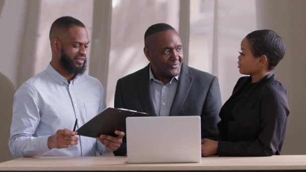 Drie collega 's collega Afrikaanse Amerikaanse vrouw, volwassen afro man baas en zwarte etnische jonge man zitten aan tafel met laptop en documenten op zoek in papieren map bespreken project praten lachen - Video