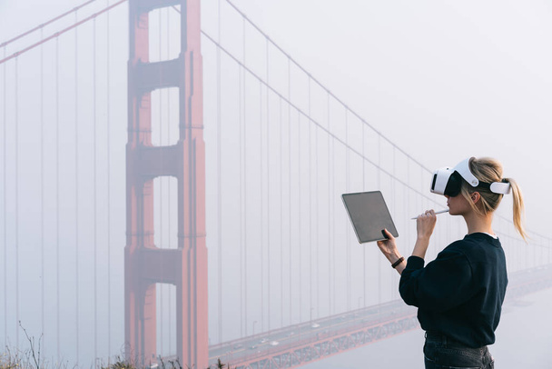 Pensive female wearing virtual reality goggles while working on tablet and planning project in headset while standing near Golden Gate in San Francisco - Fotó, kép
