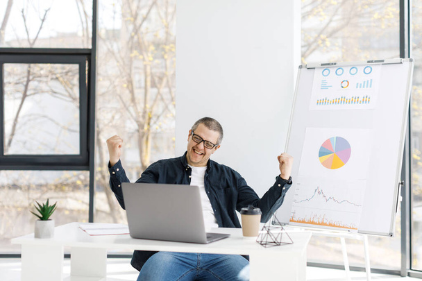 Ein Mann in legerer Kleidung arbeitet im Büro mit einem Laptop und drückt positive Emotionen aus. Der Mann arbeitet an einem abgelegenen Arbeitsplatz - Foto, Bild